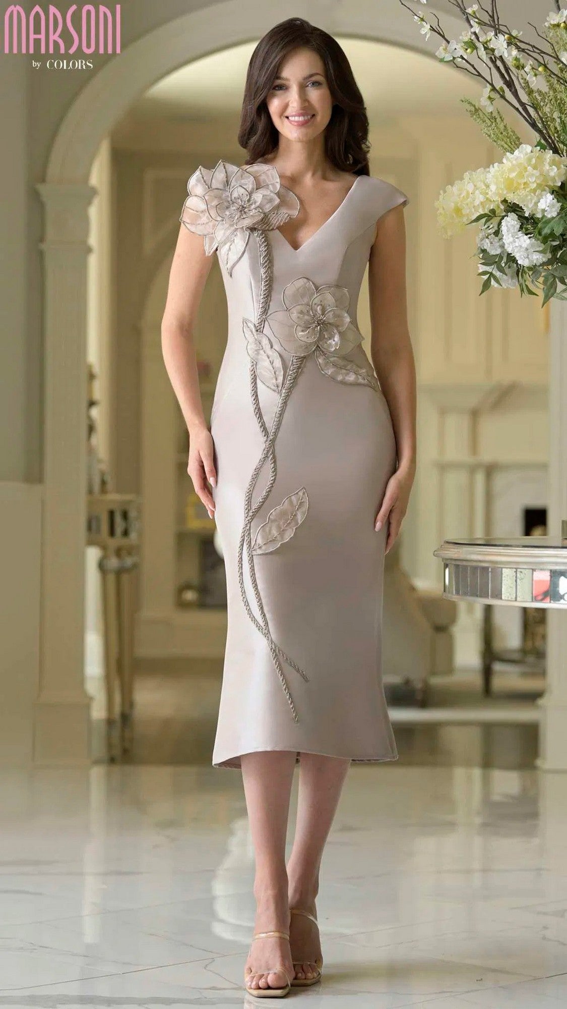 A woman stands indoors wearing the Marsoni MV1386 knee-length semi-formal dress in taupe, detailed with 3D floral appliques, and paired with nude heels.